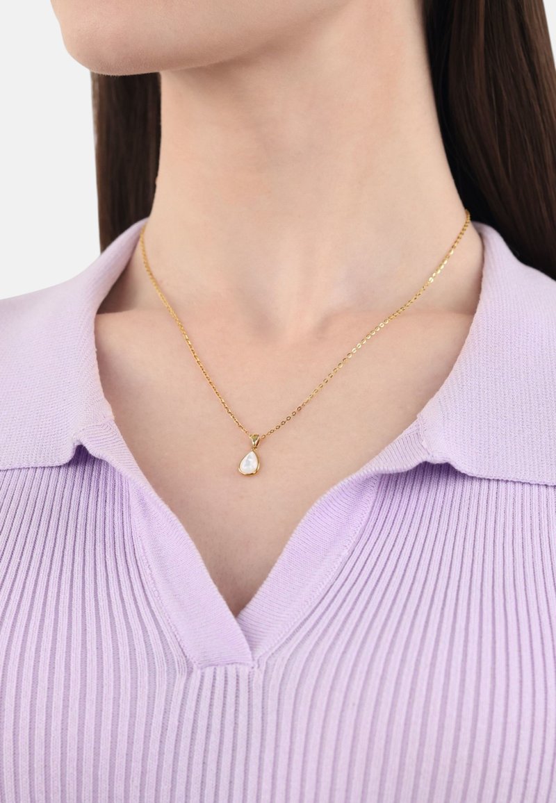 Złoty naszyjnik z wiszącym pendantem w kształcie kropli, ozdobionym białym, iryzującym kamieniem, noszony z lawendową bluzką z prążkowanym kołnierzykiem.