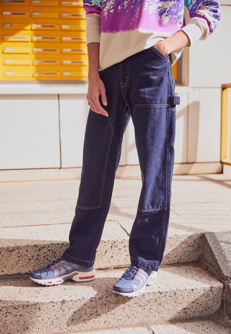 Pantalones vaqueros azul marino con costuras en contraste, corte holgado y bolsillo lateral. Combinados con zapatillas deportivas azules con detalles en rojo y diseño texturizado.