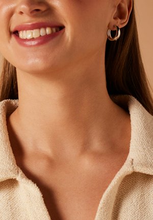 Close-up van een lachende vrouw met zilveren creooloorbellen en een beige gestructureerd overhemd met kraag.