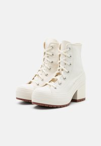 Weiße Canvas-Ankle-Boots mit einer massiven Plattform und Blockabsatz, die über Metallösen und flache Schnürsenkel verfügen. Gummi-Spitzenschutz und profiliertes Sohlenprofil.