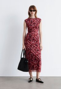 Rotes Kleid mit Blumenmuster, körperbetont, kurze Ärmel und mittellanger Saum. Kombiniert mit einer schwarzen Tragetasche und flachen Schuhen.