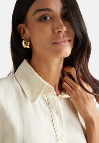 Camisa de lino color crema con cuello puntiagudo, cierre de botones y textura visible; modelo lleva pendientes de aro dorados con un aspecto natural.