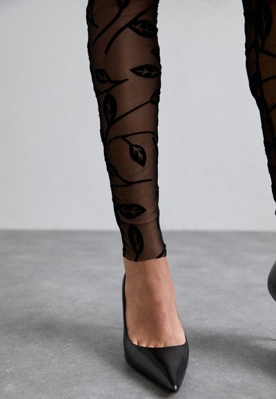Black sheer tights with leaf patterns extend to the mid-calf, paired with pointed black high heels on a gray surface.