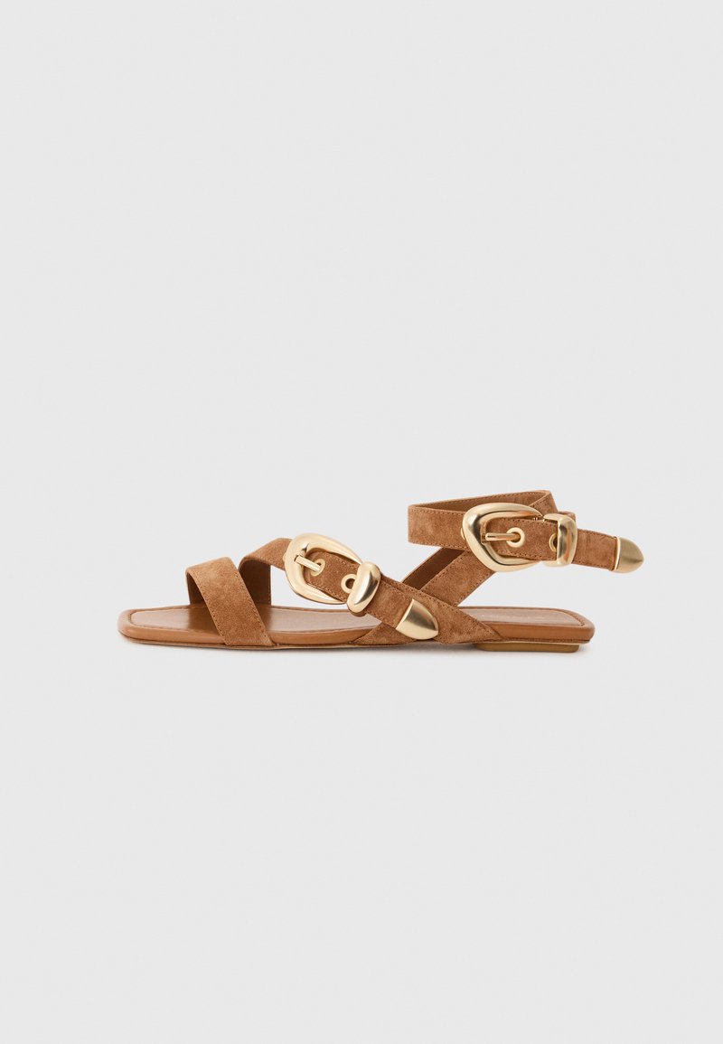 Sandales en daim marron avec des lanières entrecroisées, des accents de boucle en or et une semelle plate. Le design comporte un dos ouvert et un bout arrondi.
