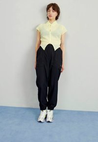 Blusa amarilla texturizada de manga corta con cuello puntiagudo, combinada con pantalones negros holgados y zapatillas blancas con acentos grises y amarillos.