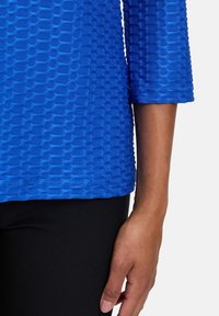 Niebieska fakturowana bluzka z rękawem 3/4, z wzorem przypominającym siatkę i gładkim wykończeniem, w połączeniu z czarnymi spodniami.