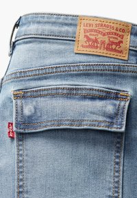 Svijetloplave traperice s smešnim kožnim detaljem, ukrašene zlatnim šavovima i crvenom Levi's oznakom na bočnoj džepu. Vidljiv dizajn poklopca džepa.