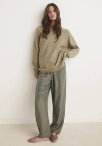Beige gebreide trui met een relaxte pasvorm en geribbelde manchetten, gedragen met losse olijfgroene broek. Zachte roze platte schoenen met groene accenten.