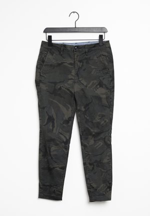Pantalones slim fit en camuflaje verde oscuro y negro con botón y cremallera, bolsillos delanteros, colgados en una percha negra sobre fondo blanco.