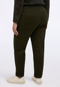 Osoba v tmavě zelených zúžených kalhotách s kapsami na zadní straně, tmavém svetru a béžových teniskách na jednobarevném bílém pozadí.