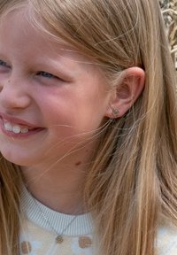 Meisje met lang, steil blond haar dat een lichtgekleurde trui met bloempatronen draagt en een zilveren oorbel in de vorm van een bloem.