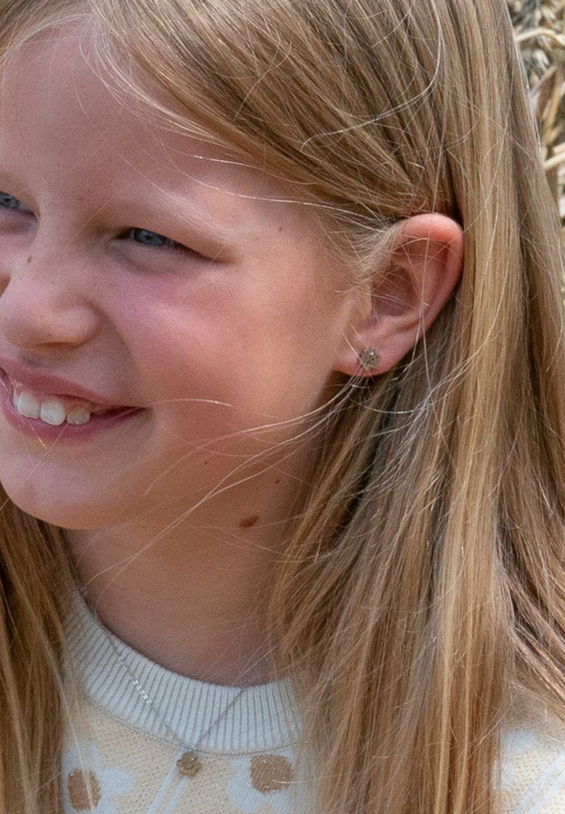 Meisje met lang, steil blond haar dat een lichtgekleurde trui met bloempatronen draagt en een zilveren oorbel in de vorm van een bloem.
