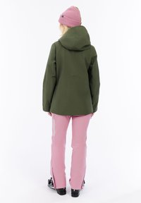 Persoon met de rug naar de camera gekeerd, gekleed in een groene hoodie, roze broek met ritszakken, een roze muts en zwarte skischoenen.