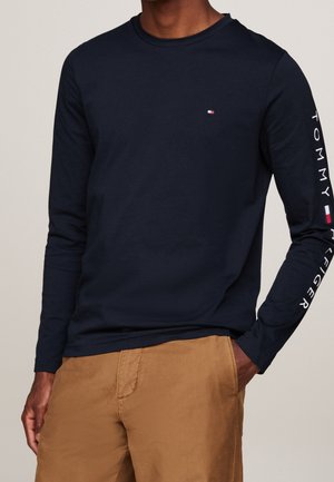 Mand iført en mørkeblå langærmet Tommy Hilfiger-skjorte med logo på brystet og højreærme, kombineret med lysebrune bukser.