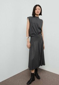 Un haut gris sans manches associé à une jupe longueur cheville assortie avec des plis, portée avec des bottes noires à lacets, sur un fond uni.