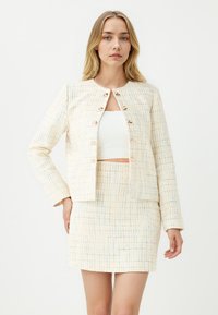 Twee-delig outfit bestaande uit een crèmekleurige tweedjas met gouden knopen en een bijpassende rok. Gelaagd stof met multicolore draden.
