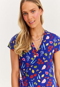 Blondýnka v pestrobarevných modrých šatech se vzory ovoce a zeleniny, s vrstvenými náhrdelníky z korálků, usmívající se s lehkým make-upem a volnými vlasy.