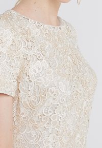 Detailní záběr na ženu, která má na sobě béžový krajkový top s květinovou výšivkou a krátkými rukávy, viditelný je krk a část její tváře a ramene.