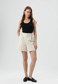 Zwart geribd mouwloos shirt, hoog uitgesneden crème korte broek met plooien en zijzakken, gecombineerd met zwarte loafers met gouden hardware accenten.