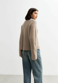 Beige geribbelde trui met hoge hals en lange mouwen, gecombineerd met lichtblauwe, losse jeans. Achtergrond is egaal en wit.
