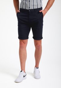 Pantalones cortos de algodón azul marino con puños doblados, que presentan bolsillos laterales y un cierre de botón. Llevados con zapatillas blancas.