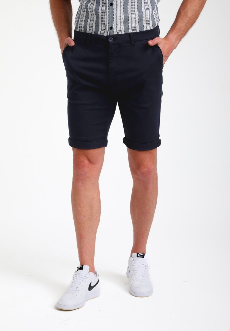 Pantalones cortos de algodón azul marino con puños doblados, que presentan bolsillos laterales y un cierre de botón. Llevados con zapatillas blancas.
