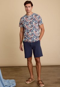 Grijs t-shirt met korte mouwen en een kleurrijk bloemenpatroon, gecombineerd met donkerblauwe shorts en bruine sandalen met een bandontwerp.