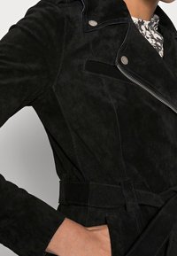 Svart mockajacka med figursydd design, dragkedja och bälte i midjan. Den har silverfärgade detaljer och en slät textur.