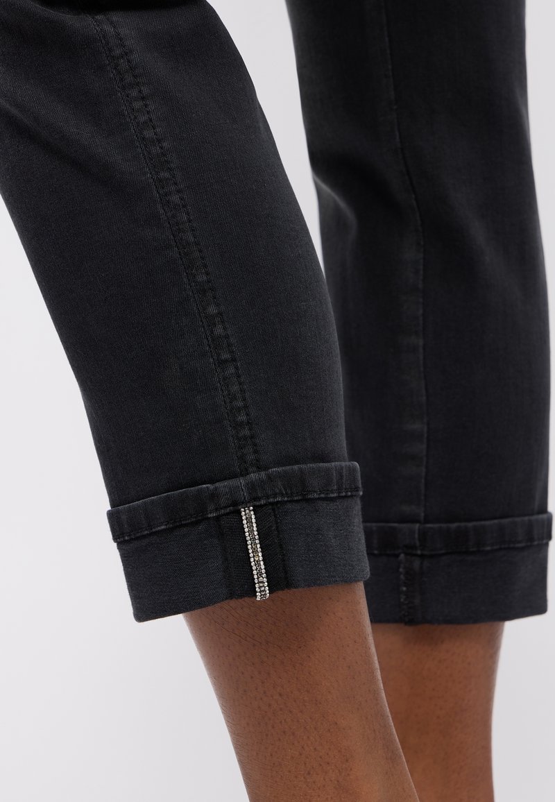 Gros plan sur un jean noir retroussé avec une petite rangée verticale de strass sur le revers extérieur, porté sur une peau foncée.