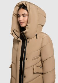Beezi puffer jakk, millel on suur kapuuts, lukuga eesmine osa ja kaks eesmist taskut. Omab niidiga õmmeldud polsterdust ja sileda tekstuuri.