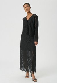 Schwarzes Spitzenkleid mit langen Ärmeln, einem V-Ausschnitt mit Bindung und einem durchsichtigen, bodenlangen Design mit gewelltem Saum.