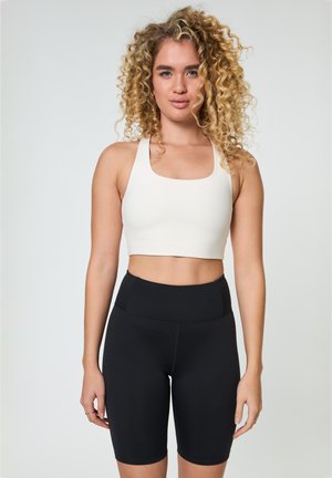 Kvinne med krøllete blondt hår iført hvit croptopp og høytlivs svarte sykkelshorts, stående foran en ensfarget bakgrunn.