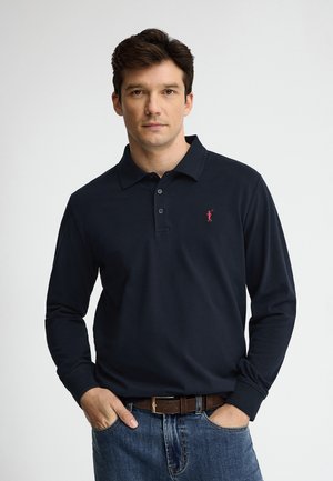 Homme aux cheveux foncés portant un polo à manches longues bleu marine avec un logo rouge et un jean bleu, mains dans les poches avant, fond neutre.