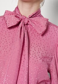 Rosa blus i textil med struktur och en stor rosett vid kragen. Har en subtilt mönstrad design med små, glänsande detaljer genom hela plagget.