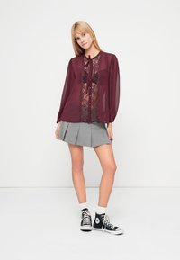 Burgundia läbipaistev pluus pitsist esiküljega ja nööpidega, kombineeritud volditud halli seelikuga, valgete sokkide ja mustade kõrgete tennistega.