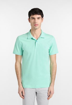 Jonge man met kort donker haar, gekleed in een lichtgroene korte mouw polo en lichtgrijze broek, staand tegen een effen witte achtergrond.