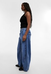 Femme aux longues tresses portant un haut noir sans manches, un jean bleu à jambes larges et des chaussures noires, debout de profil sur un fond blanc.