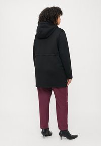 Manteau à capuche noir avec une coupe décontractée, doté d'une fermeture éclair et d'une couture horizontale dans le dos. Associé à un pantalon bordeaux et des bottines.