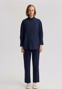 Camisa de botones de color azul marino con mangas largas, combinada con pantalones a juego. Ambos elementos presentan una textura suave y un corte relajado, usados con zapatos beige.