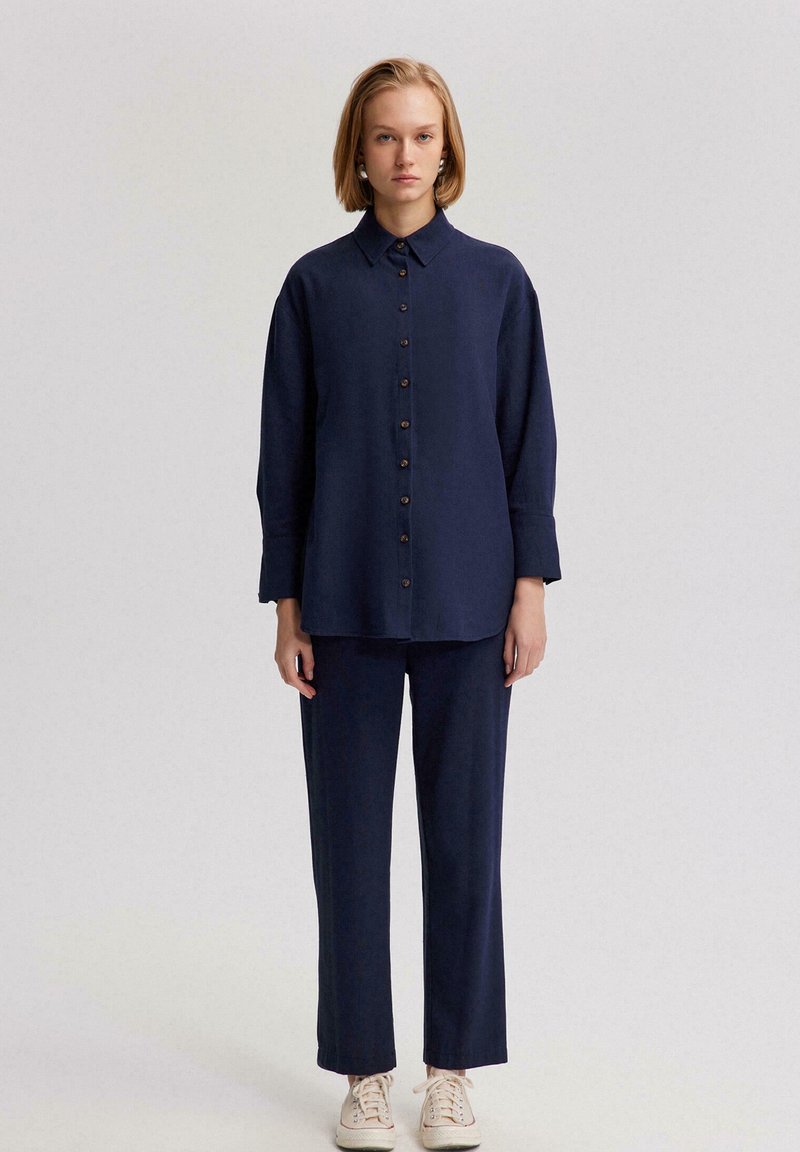 Camisa de botones de color azul marino con mangas largas, combinada con pantalones a juego. Ambos elementos presentan una textura suave y un corte relajado, usados con zapatos beige.