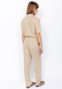 Jumpsuit beige in lino con maniche corte, piega sul retro e due tasche posteriori; presenta una vestibilità morbida e polsini delle maniche arrotolati.