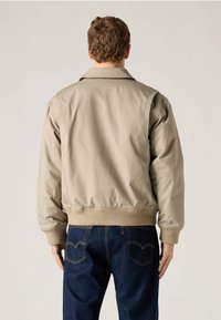 Άνδρας με μπεζ μπουφάν bomber και σκούρο μπλε τζιν, στέκεται πλάτη σε απλό ανοιχτό φόντο.