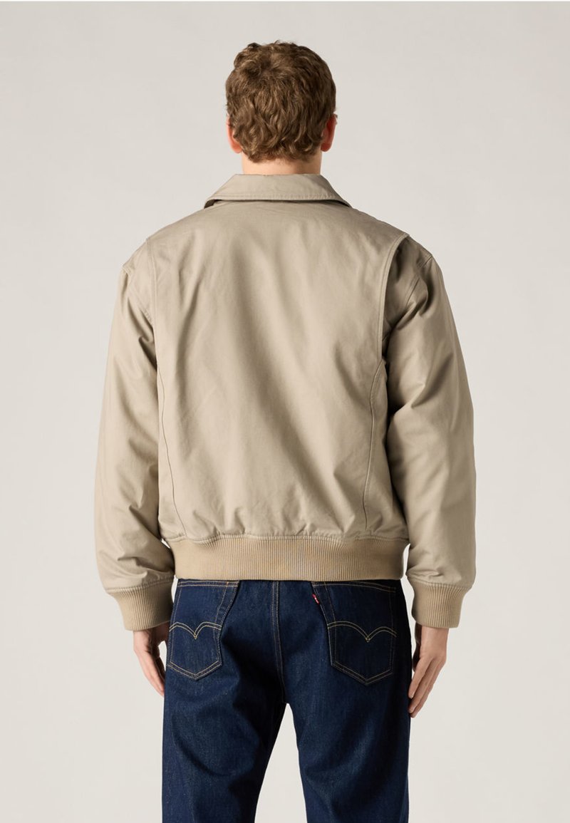 Uomo con giacca bomber beige e jeans blu scuro, in piedi di spalle su sfondo chiaro uniforme.
