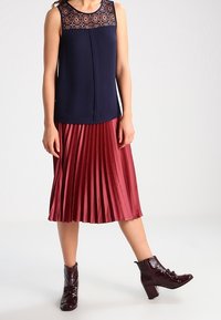 Námorně modrá tílko bez rukávů s krajkovými detaily a plisé sukně v burgundské barvě do midi délky, doplněná o lesklé burgundské kotníkové boty na blokovém podpatku.