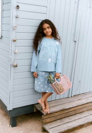 Cămașă albastru deschis cu mâneci lungi și text alb, fustă din denim cu modele de inimă, și coș roz din plastic care ține un ananas, desculț pe scări de lemn.