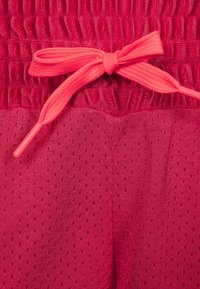 Pantaloni sportivi rossi con una vita elasticizzata arricciata e un laccio di chiusura rosa brillante. Il tessuto presenta una texture traspirante e perforata.