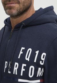 Sudadera con cremallera de color navy, fabricada en una mezcla de algodón, que presenta el texto blanco "FQ19 PERFORM" y un detalle de bloque de color horizontal. Diseño texturizado y casual.