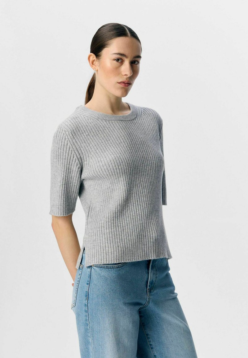 Maglione grigio a coste con maniche corte, scollatura rotonda, fessure laterali e texture morbida, abbinato a jeans blu a vestibilità rilassata.