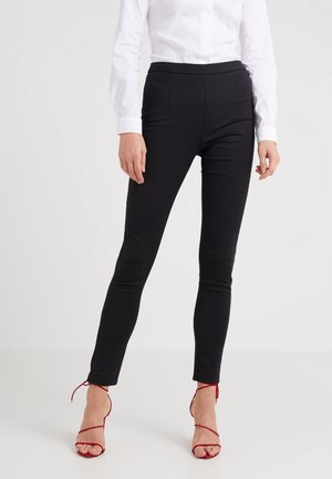 Mujer con pantalones negros ajustados, camisa blanca de manga larga y sandalias rojas de tacón alto con tiras, de pie sobre un suelo blanco.