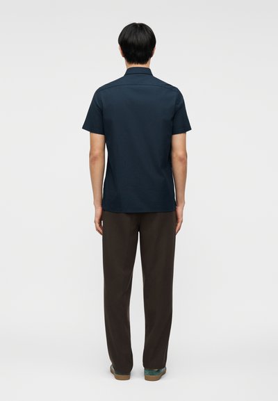 Άνδρας που στέκεται με την πλάτη, φοράει σκούρο μπλε κοντομάνικο πουκάμισο με γιακά, σκούρα καφέ παντελόνια και casual παπούτσια σε απλό φόντο.